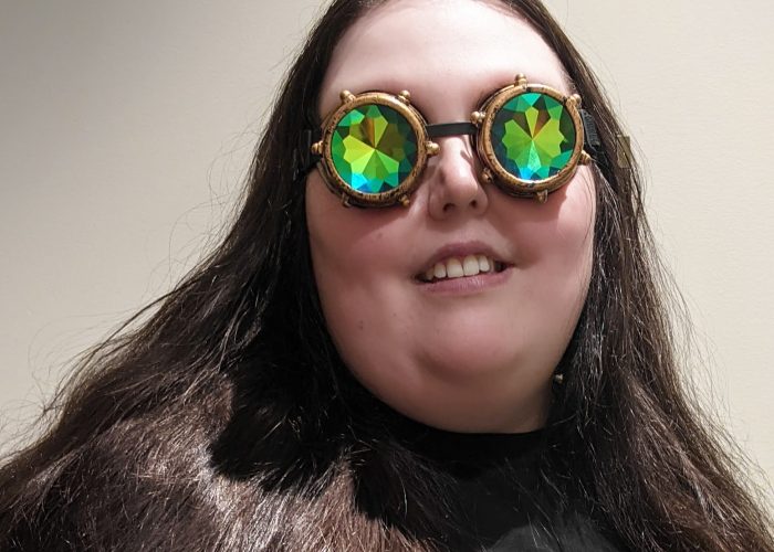 Headshot of Susan, a Fat, Pākeha person with long brown hair. They are smiling at the camera and wearing a pair of sparkly-reflective goggles in the fashion style known as Steampunk.