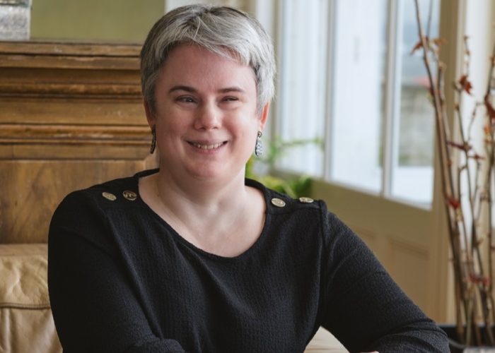 A head and shoulders shot of Lauren sitting on a beige couch. She is smiling, and has short-cropped, grey hair. She is wearing a black top with button details on the shoulders. The background includes a wooden cabinet and decorative plants near a window. 